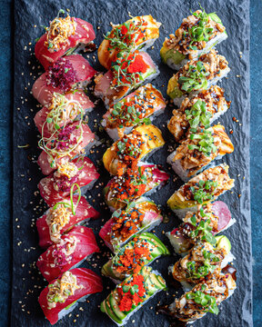 Colorful assorted sushi rolls arranged in rows on a dark slate platter, garnished with fresh herbs, sesame seeds, and sauces.