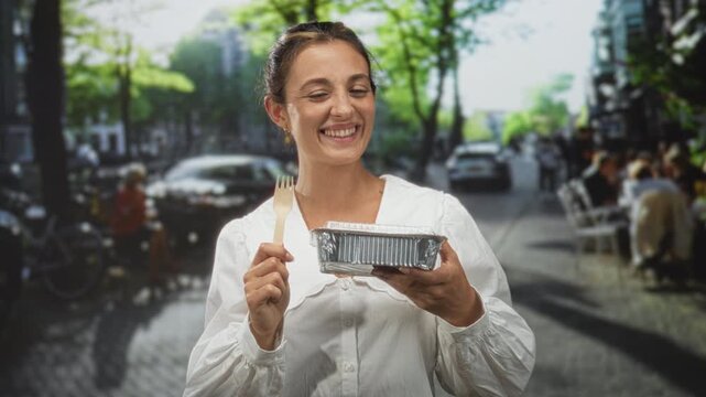 Woman smiling holds takeout container and wooden fork on city street in daylight; casual community dining joy.