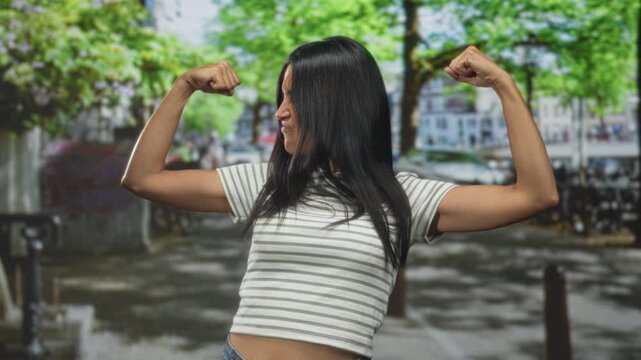 Woman flexing biceps with fists clenched, striped cropped top showing midriff on a leafy urban street with parked bikes; confidence empowerment.