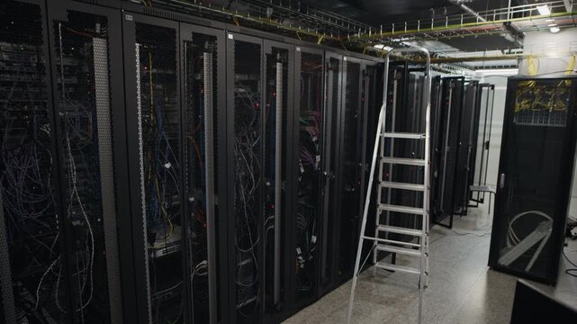 Long aisle of server racks with visible network cables and maintenance ladder in modern data center facility, wide static shot showing cloud computing infrastructure and server hardware environment