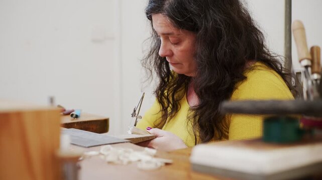 Focused female jeweler cutting silver metal with a piercing saw at her workbench