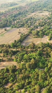 Aerial shot of lush green fields and scattered trees in the countryside near Banteay Samre, Cambodia. The landscape features agricultural plots and patches of forest in warm sunlight.