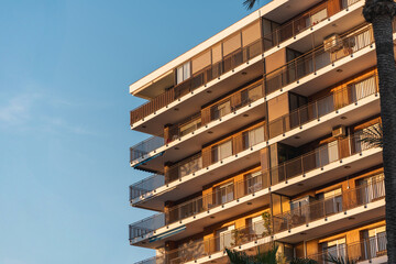 Obraz premium Side view of a modern apartment building with repeating balconies lit by warm sunset light against clear blue sky, illustrating urban living, housing, and real estate concepts.