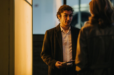 A male business associate speaks to a female colleague while holding a clipboard in an evening...