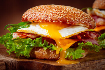 Two juicy burgers sit on a wooden board, each filled with lettuce, bacon, cheese, and fried eggs.