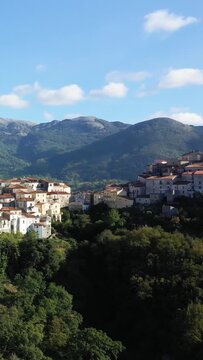 Picturesque view of Aieta village nestled on a lush green hillside with mountains in the background under a blue sky. Traditional Mediterranean architecture with red-tiled roofs stands out against the