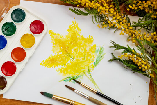 A bouquet of mimosa flowers painted in watercolor next to paints and a branch of yellow flowers. Student art at school. 