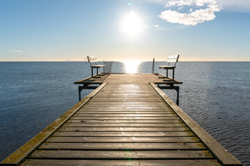 A straight wooden pier leads toward open water and a bright sunlit horizon, with simple benches at the end. Minimal coastal scene ideal for relaxation, contemplation, travel, freedom and peaceful seas © Volha