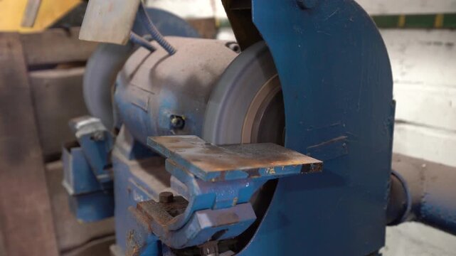 A worker operates a grinding machine in a workshop. The machine grinds a metal piece for sharpening. The light comes from the nearby window. Dust fills the air as the work continues.