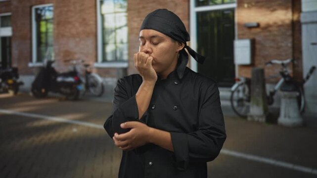 Man chef in black uniform with hand to chin on street near brick building and parked bicycles; quiet thinking.