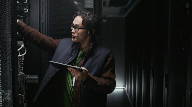 Indoor medium slowmo shot of Asian male network technician standing by opened server rack checking cables and ports while reviewing on tablet and communicating via headset in dark data center