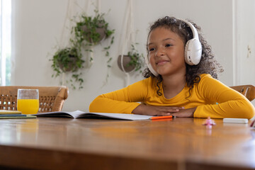 African American girl child sitting at home dining table with white headphones, open notebook © wavebreak3