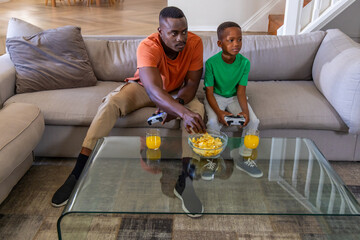 African American father and son gaming on light-gray sofa with controllers, chips and juice © wavebreak3