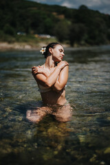Fototapeta premium A woman stands in clear river water, arms crossed over her chest, eyes closed. The sunlit scene captures a moment of calm and natural beauty by the river.
