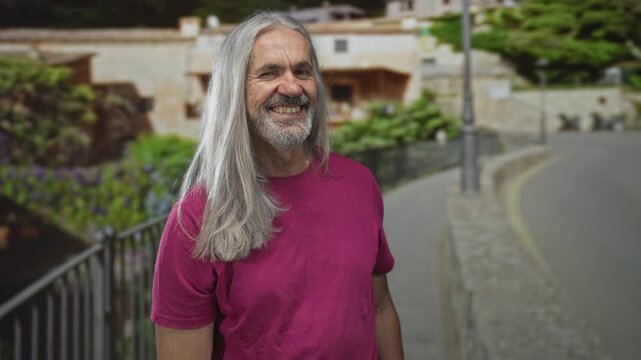 Man with long silver hair smiling broadly showing teeth on a street beside stone railing; carefree joy.