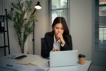 Young Asian businesswoman sitting at desk, deeply thinking with hand on chin, feeling stressed