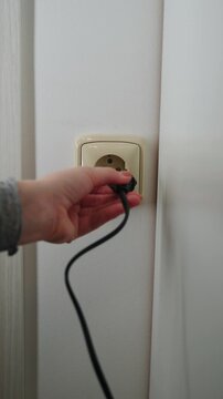 Close-up of hand trying to plug electrical cord into wall outlet, but plug doesn&rsquo;t fit, showing electrical installation and household interior.