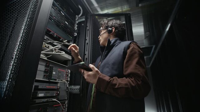 Indoor low angle shot of Asian male technician installing server rack equipment and connecting cables while checking on digital tablet and communicating through headset in data center