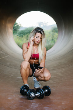 A motivated fitness athlete taking a break to play music on her smartphone. Female athlete resting during outdoor workout with dumbbells