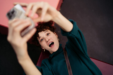 Excited red-haired girl making surprised face while snapping selfie outdoors. Whimsical expression...