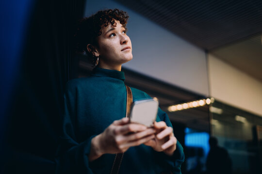 Freelancer with smartphone looks upward thoughtfully, symbolizing creative vision, tech-driven purpose, and emotional connection to mobile work in modern digital landscape.