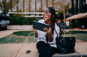 Obraz premium Laughing woman with vitiligo sitting outdoors with tablet, successful freelance work and positive lifestyle, authentic diversity in tech and business concept
