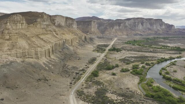 Aerial view of a white van driving on a dirt road through rocky cliffs and a winding river in the Patagonian arid landscape