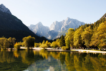 Fototapeta premium Golden autumn foliage and green pine trees reflect perfectly in the calm, clear waters of Lake Jasna in Slovenia. High quality professional stock photo