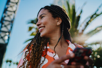 Young woman with braided hair smiling while holding digital tablet outdoors, representing creativity, empowerment, and the freedom of modern mobile technology in daily life. © BullRun