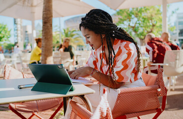 Concentrated woman leaning forward and writing in notebook with tablet nearby, representing...