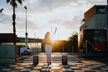 Woman wearing headphones walks across crosswalk at sunset, hand raised in the air, embodying empowerment, digital freedom, creativity and lifestyle expression. © BullRun