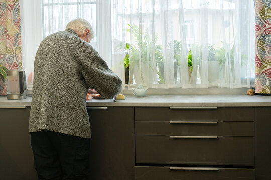From behind, an elderly man stands at kitchen counter preparing food. The view emphasizes his posture and quiet focus of daily chores. Grandpa engaged in cooking.