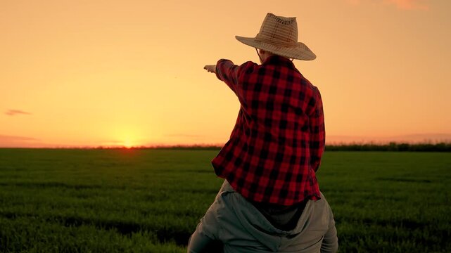 Child boy play together father in green field. Dad son point toward sunset. Happy family. Kid boy in hat sits on father's shoulders. Family trip through nature park. Child shows dad sunset with hand