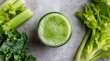 Obraz premium Fresh green juice in clear glass jar surrounded by celery stalks and leafy greens on a textured gray surface, top view with natural lighting