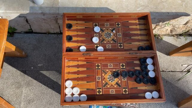 Backgammon game with wooden board and black and white pieces outdoors casting shadows
