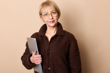 Naklejka premium Mature woman with glasses wearing a brown jacket holds a laptop and looks at the camera. Neutral background with soft lighting creates a calm professional atmosphere.