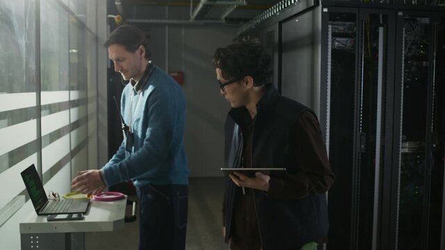 Two male network engineers using digital tablet and laptop and communicating while walking along data center corridor and entering server rack, tracking eyelevel medium shot