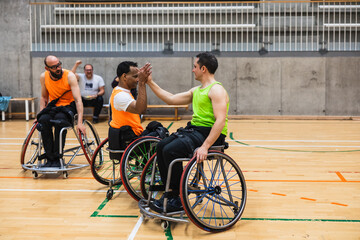 Two diverse men in sport wheelchairs high fiving on a gymnasium court, celebrating teamwork and success during a basketball match © Jordi Mora