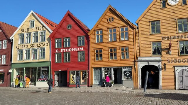 People walk along the waterfront in Bergen, Norway. Brightly colored buildings line the street. The weather is clear with a blue sky. People enjoy their time outside.