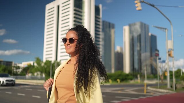 Young Brazilian businesswoman with sunglasses crossing urban street in summer sunlight with modern skyscrapers