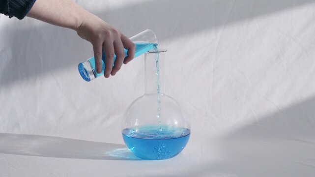 pouring blue chemical solution from a graduated cylinder into a round bottom flask
