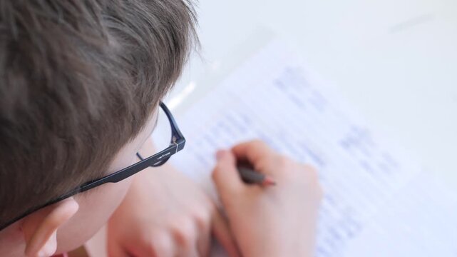 Student boy wearing glasses concentrating intently on writing notes and completing homework on a paper sheet with a pencil, focusing on education and learning