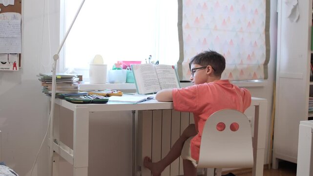 Young boy with glasses sits at a white desk in his room, focused on reading a textbook and doing homeworkconcentrating on learning and schoolwork at home during daytime