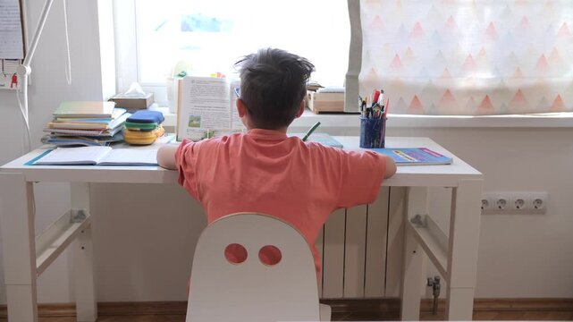 Young boy sits at white desk studying an open textbook with school supplies around, concentrating on homework in a sunlit room focused home learning and childhood diligence