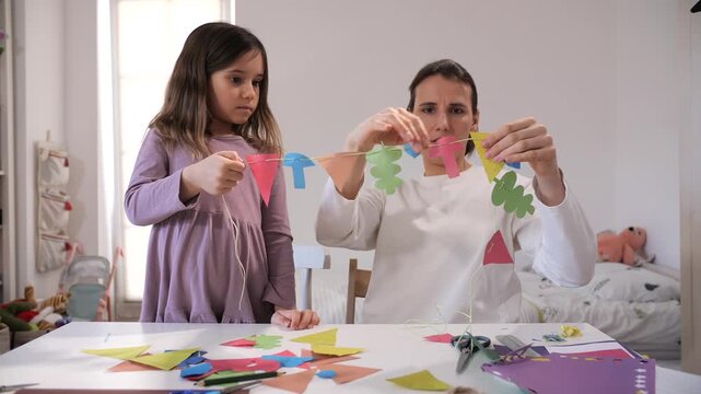 Mother and daughter bonding crafting at a white table, meticulously threading colorful paper cutouts onto a string, involving teamwork and focused attention in a bright room