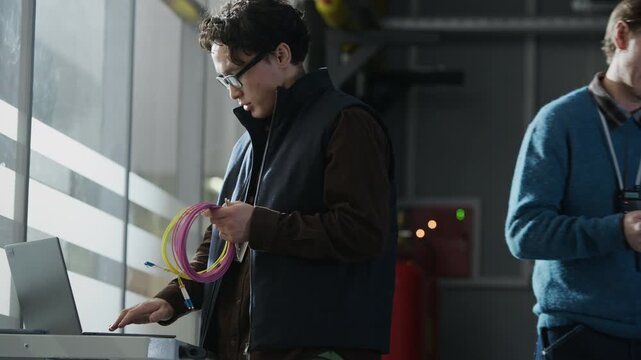 Side view medium shot of Asian male network engineer holding fiber optic cable coil while operating laptop on workstation cart in modern data center corridor