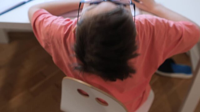 Young boy wearing glasses and a pink t shirt, lying upside down on a desk with a notebook, looking up playfully while studying or taking a break from homework