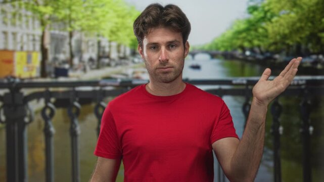 Man with hand covering face in a facepalm and shrug pose on a street by an amsterdam canal bridge, red shirt visible; frustration travel.