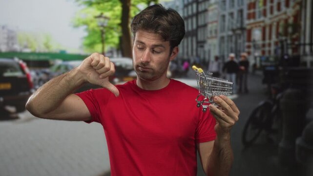 Man in red shirt holding miniature shopping cart and showing thumbs down on a street; consumer dissatisfaction disappointment.