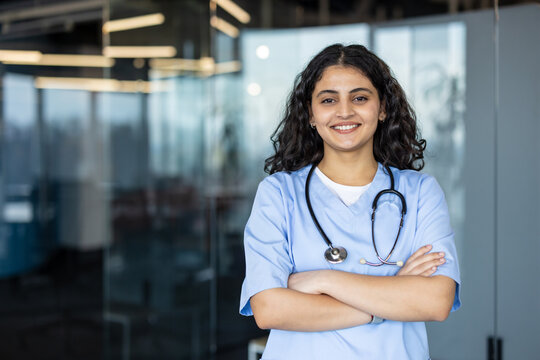 Young female medical professional in scrubs and stethoscope smiling, standing with arms crossed at a modern clinic or hospital, representing healthcare, wellness, and dedicated service